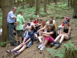 Viele Kinder sitzen auf den Waldboden und hören ihren Führer gespannt zu.